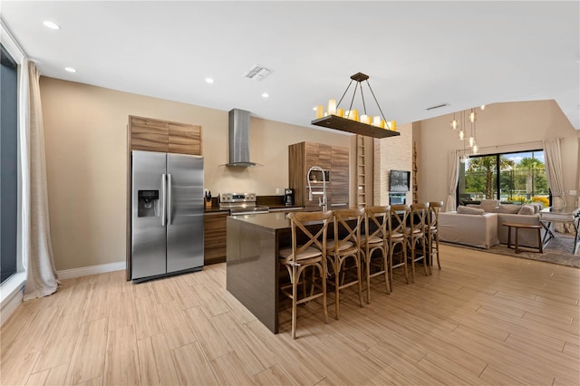 kitchen with an island with sink, a kitchen breakfast bar, appliances with stainless steel finishes, wall chimney range hood, and a notable chandelier