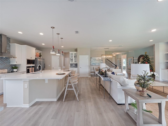 kitchen with appliances with stainless steel finishes, light hardwood / wood-style flooring, a center island with sink, wall chimney range hood, and a notable chandelier