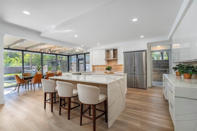 kitchen with built in refrigerator, plenty of natural light, white cabinets, and wall chimney range hood