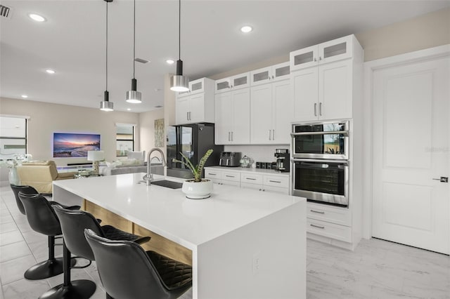 kitchen with an island with sink, pendant lighting, a breakfast bar area, double oven, and white cabinetry