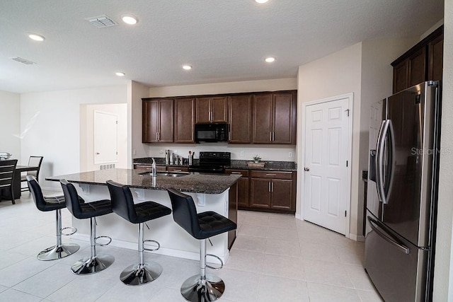 kitchen with a breakfast bar, sink, black appliances, a center island with sink, and dark stone countertops