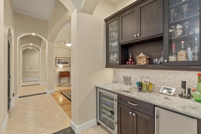 bar with light tile patterned floors, dark brown cabinets, beverage cooler, light stone countertops, and crown molding