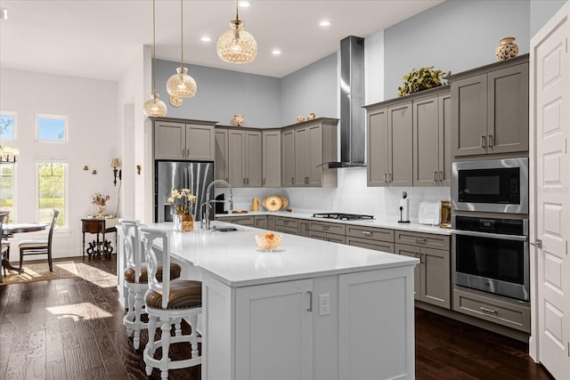 kitchen with stainless steel appliances, a kitchen island with sink, decorative light fixtures, wall chimney range hood, and sink