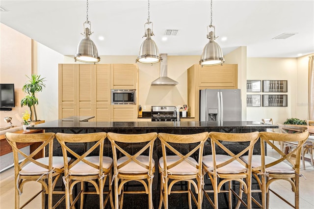 kitchen with an island with sink, a kitchen bar, light brown cabinetry, wall chimney exhaust hood, and appliances with stainless steel finishes