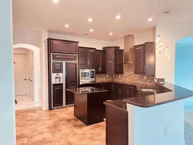 kitchen with arched walkways, a center island, built in appliances, wall chimney range hood, and a sink