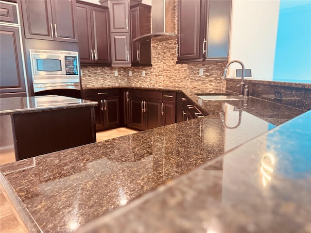 kitchen with dark stone countertops, wall chimney exhaust hood, stainless steel microwave, and a sink