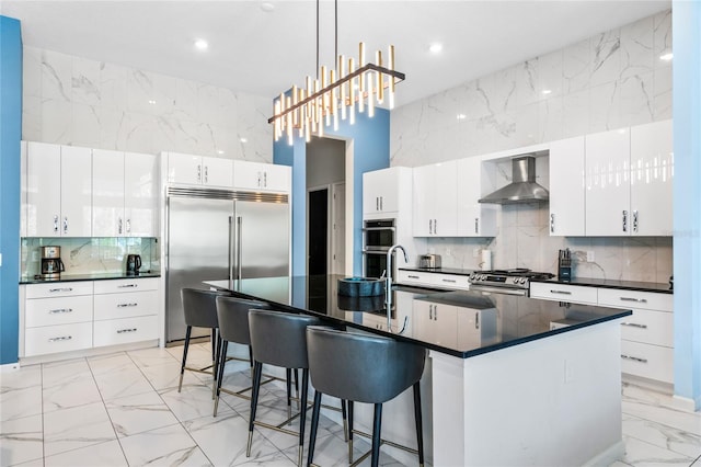 kitchen featuring white cabinetry, wall chimney exhaust hood, hanging light fixtures, stainless steel appliances, and a center island with sink