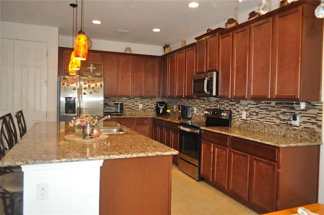 kitchen featuring light stone counters, hanging light fixtures, an island with sink, stainless steel appliances, and backsplash