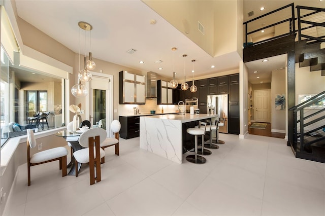 kitchen with a center island with sink, wall chimney range hood, pendant lighting, and stainless steel fridge with ice dispenser