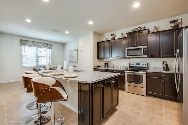 kitchen featuring light tile floors, appliances with stainless steel finishes, a center island with sink, light stone countertops, and dark brown cabinetry