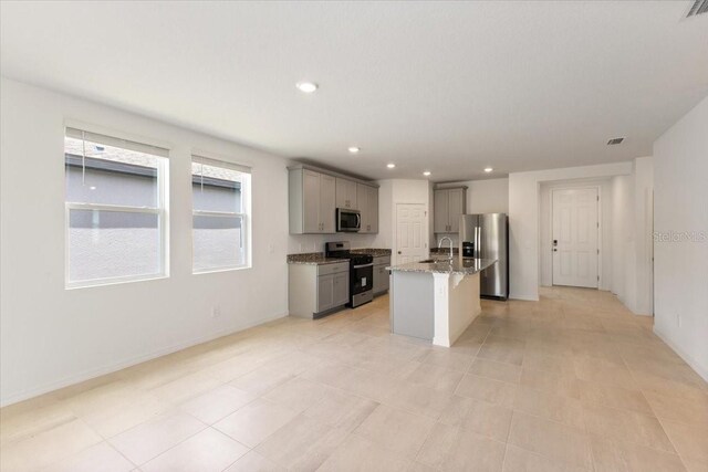 kitchen with light tile floors, gray cabinetry, stainless steel appliances, a center island with sink, and light stone countertops