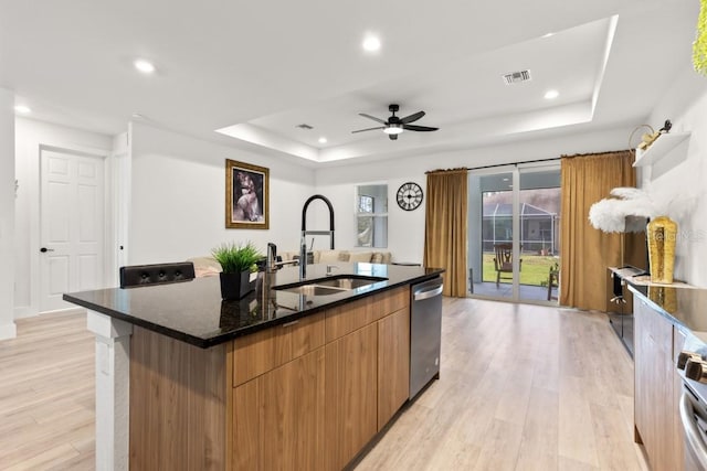 kitchen featuring sink, ceiling fan, a center island with sink, a tray ceiling, and light wood-type flooring