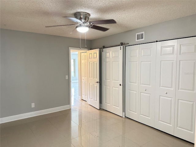 unfurnished bedroom with ceiling fan, a textured ceiling, a barn door, light tile patterned flooring, and a closet