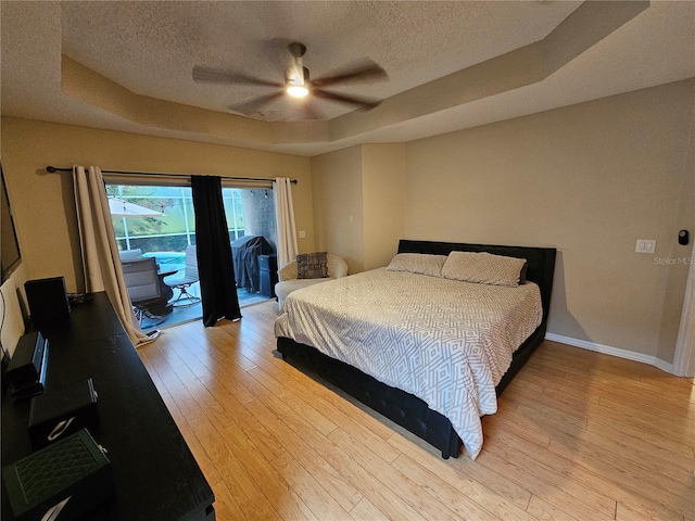 bedroom featuring ceiling fan, light wood-type flooring, access to outside, and a tray ceiling