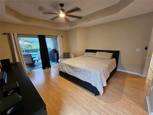 bedroom with access to exterior, light hardwood / wood-style floors, ceiling fan, and a tray ceiling