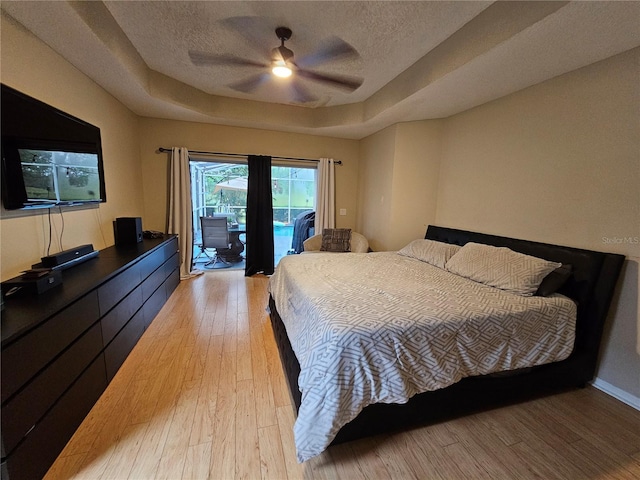 bedroom featuring access to outside, light hardwood / wood-style flooring, ceiling fan, a textured ceiling, and a tray ceiling