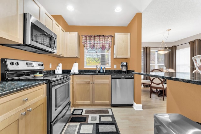 kitchen with dark stone counters, hanging light fixtures, stainless steel appliances, light brown cabinets, and light hardwood / wood-style flooring