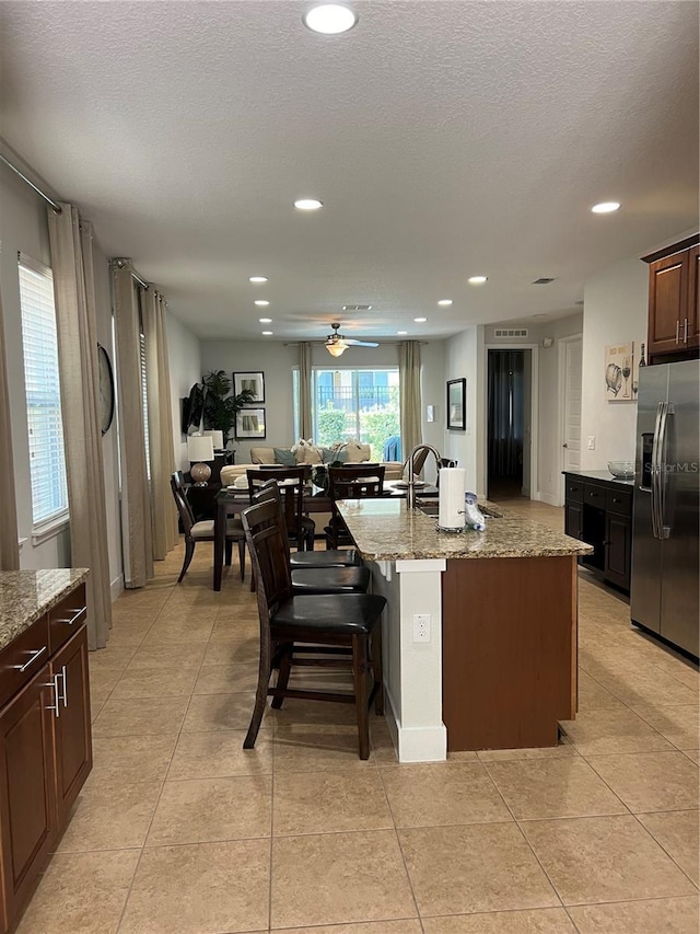 kitchen with stainless steel fridge, a center island with sink, light stone counters, open floor plan, and a breakfast bar area