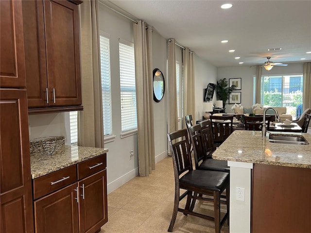 kitchen featuring a center island with sink, ceiling fan, light stone counters, a kitchen bar, and a sink