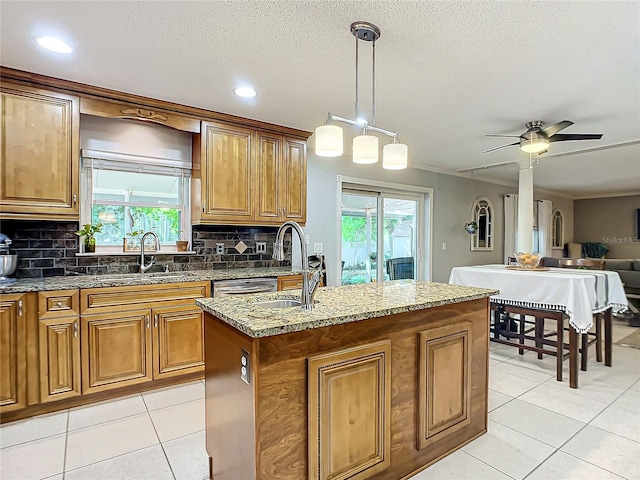 kitchen with hanging light fixtures, backsplash, ceiling fan, a kitchen island with sink, and light stone counters