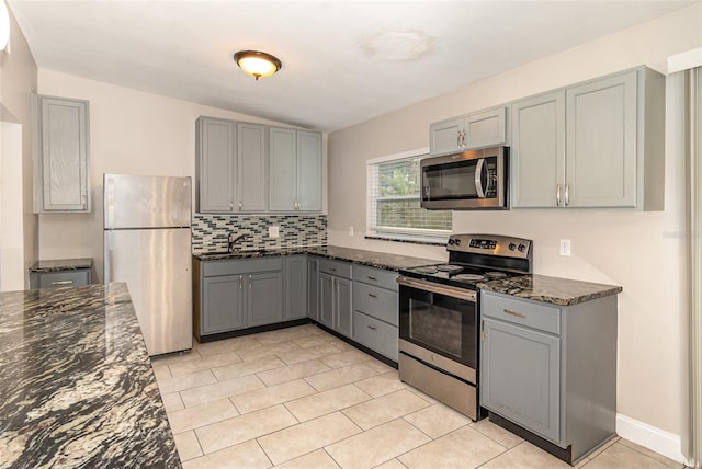 kitchen with light tile floors, dark stone counters, appliances with stainless steel finishes, gray cabinetry, and backsplash