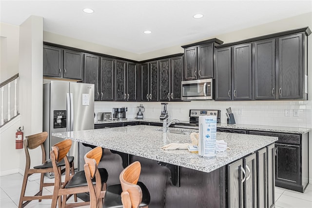 kitchen with light tile floors, appliances with stainless steel finishes, light stone counters, and a center island with sink