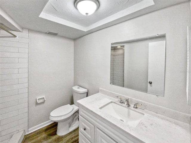 bathroom with hardwood / wood-style floors, tiled shower, a tray ceiling, toilet, and vanity