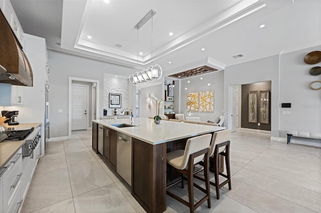 kitchen featuring an island with sink, sink, light tile floors, a raised ceiling, and white cabinets