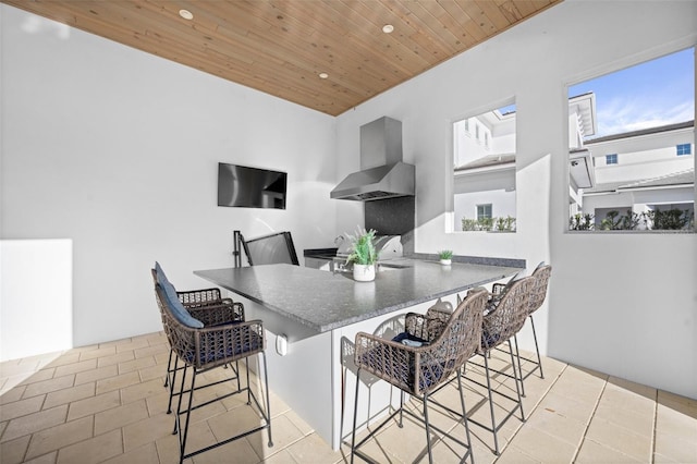 kitchen featuring kitchen peninsula, light tile floors, wood ceiling, a breakfast bar, and wall chimney exhaust hood