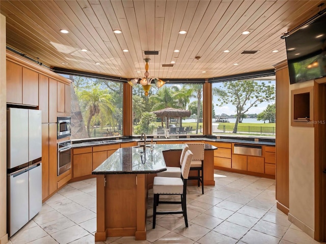 kitchen with wood ceiling, a kitchen island with sink, appliances with stainless steel finishes, dark stone counters, and plenty of natural light