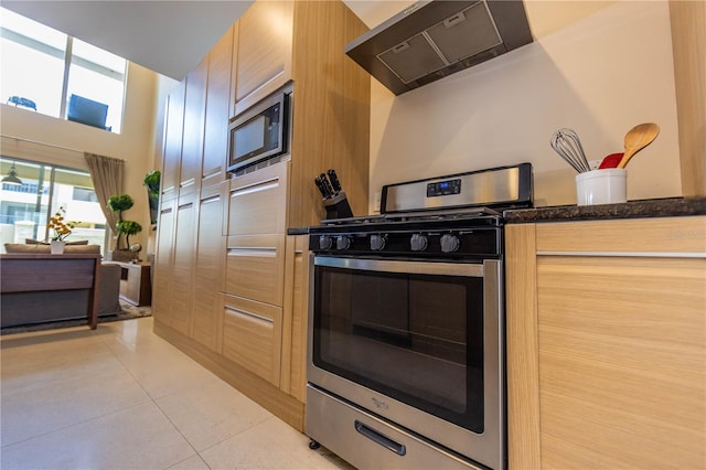 kitchen featuring appliances with stainless steel finishes, light brown cabinets, wall chimney exhaust hood, light tile floors, and dark stone countertops