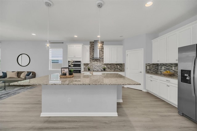 kitchen with white cabinets, hanging light fixtures, stainless steel appliances, wall chimney exhaust hood, and a center island with sink