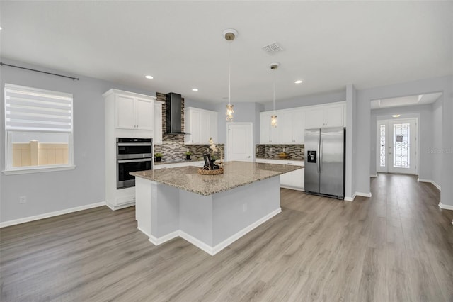 kitchen featuring light hardwood / wood-style flooring, pendant lighting, appliances with stainless steel finishes, wall chimney range hood, and white cabinets