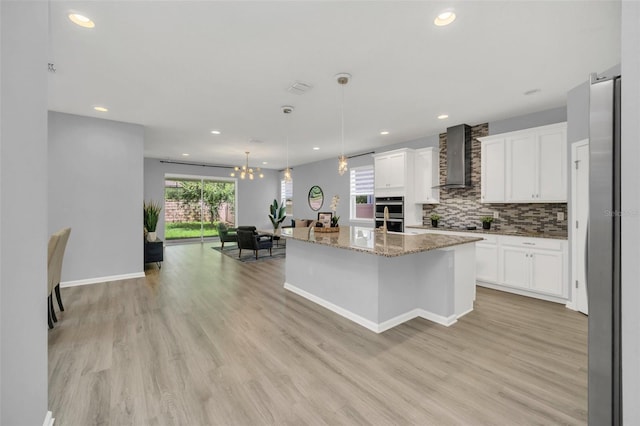 kitchen featuring white cabinetry, light hardwood / wood-style flooring, an island with sink, light stone countertops, and wall chimney range hood