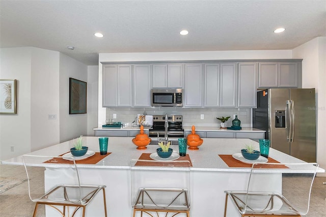 kitchen with gray cabinetry, backsplash, a center island with sink, a kitchen breakfast bar, and appliances with stainless steel finishes