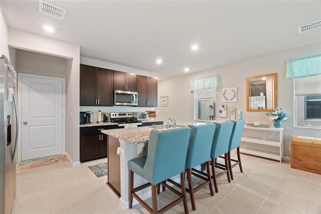 kitchen featuring light stone countertops, a breakfast bar, stainless steel appliances, a center island with sink, and dark brown cabinetry