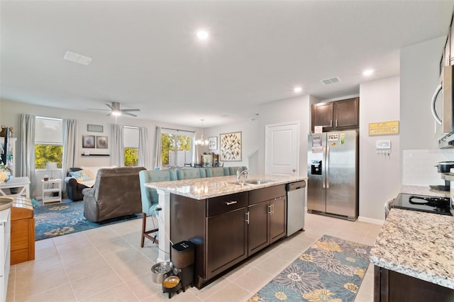 kitchen featuring an island with sink, stainless steel appliances, ceiling fan with notable chandelier, dark brown cabinets, and sink