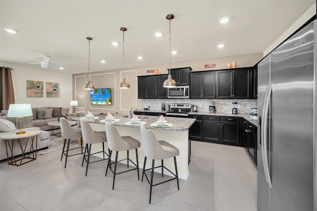 kitchen featuring appliances with stainless steel finishes, a breakfast bar, decorative light fixtures, light stone counters, and a center island with sink