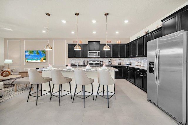 kitchen featuring a kitchen bar, hanging light fixtures, appliances with stainless steel finishes, an island with sink, and light stone countertops