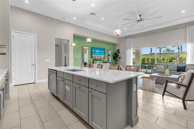 kitchen with ceiling fan, sink, a tray ceiling, an island with sink, and light stone countertops