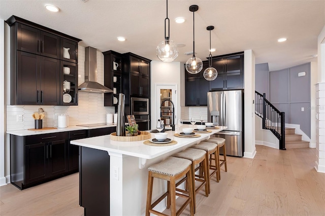 kitchen featuring stainless steel refrigerator with ice dispenser, backsplash, wall chimney exhaust hood, a center island with sink, and decorative light fixtures