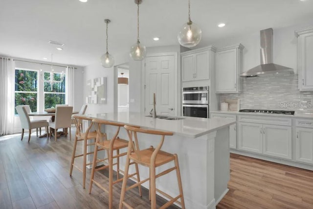 kitchen with backsplash, stainless steel appliances, wall chimney range hood, white cabinets, and light wood-type flooring