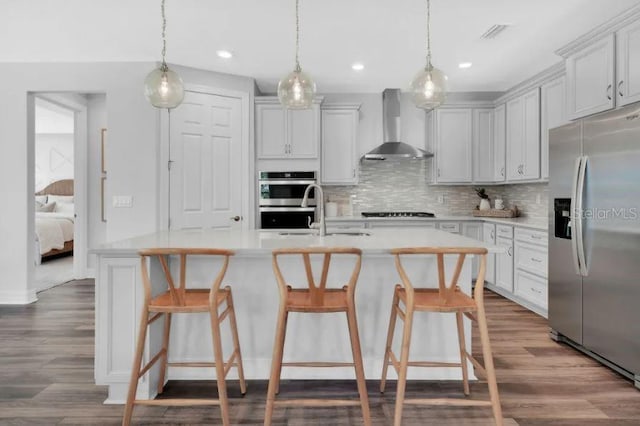 kitchen featuring appliances with stainless steel finishes, wall chimney range hood, backsplash, hardwood / wood-style flooring, and a kitchen island with sink