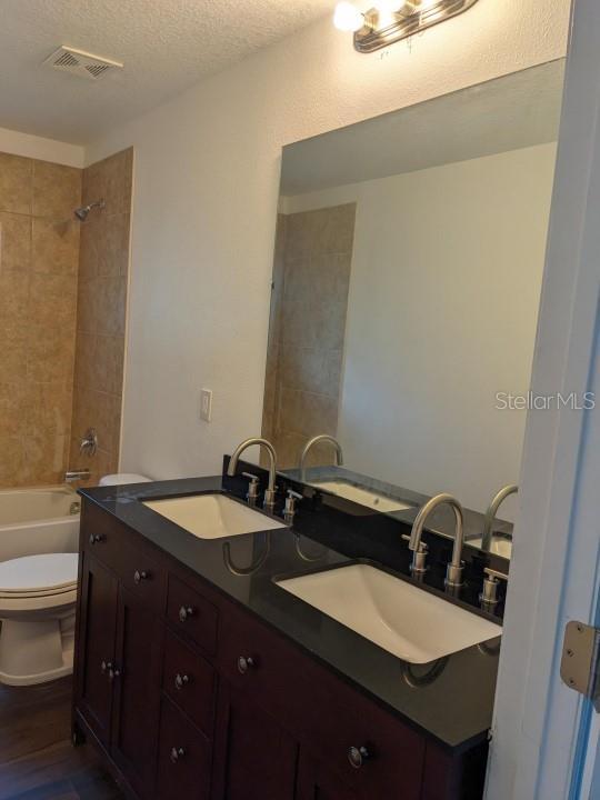 full bathroom featuring a textured ceiling, dual bowl vanity, tiled shower / bath combo, toilet, and wood-type flooring