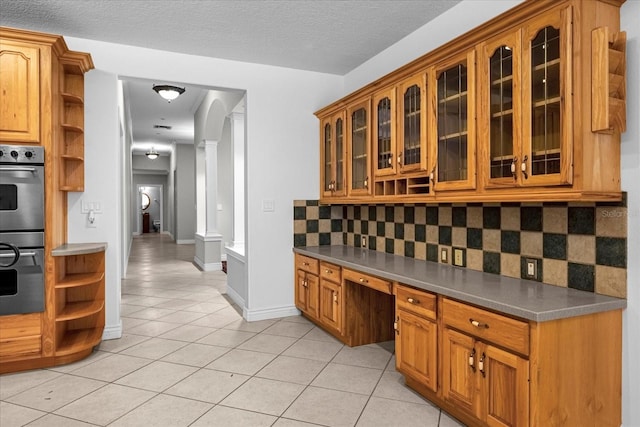 kitchen with double oven, light tile floors, tasteful backsplash, ornate columns, and a textured ceiling