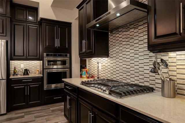 kitchen with wall chimney exhaust hood, light stone countertops, stainless steel appliances, tasteful backsplash, and dark brown cabinetry