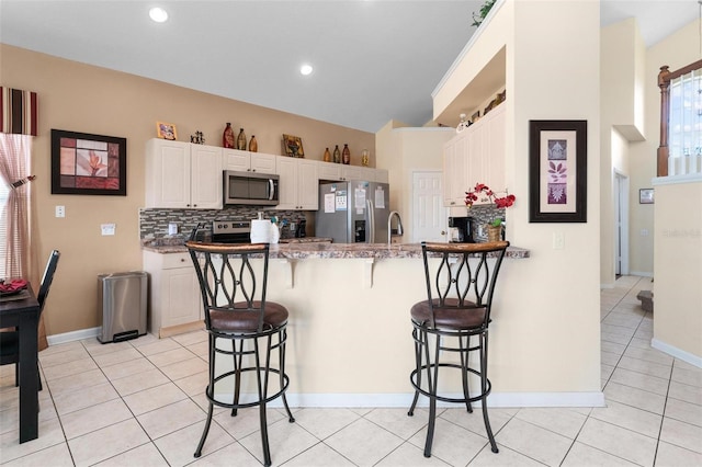 kitchen with appliances with stainless steel finishes, backsplash, a breakfast bar area, light tile flooring, and white cabinetry