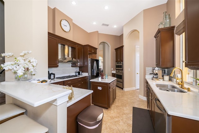 kitchen with wall chimney exhaust hood, kitchen peninsula, appliances with stainless steel finishes, sink, and tasteful backsplash