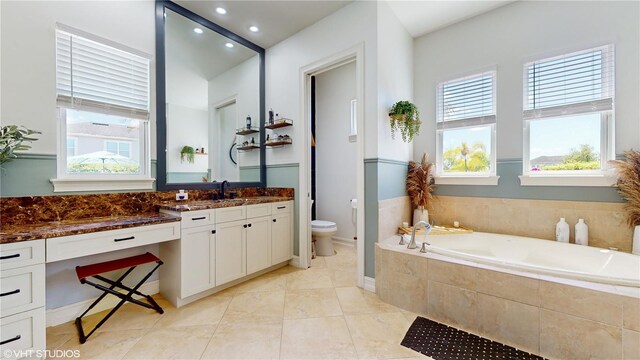 bathroom with a wealth of natural light, vanity, toilet, and tiled bath