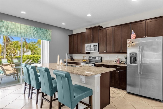 kitchen featuring a kitchen island with sink, light stone countertops, light tile flooring, appliances with stainless steel finishes, and sink